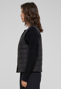 Doudoune noire sans manches avec un design matelassé, dotée d'un col rond, portée par-dessus un pull noir à manches longues.