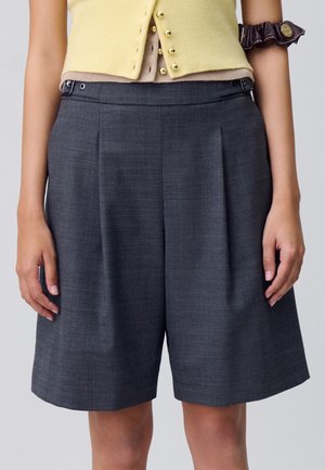 Shorts plissés gris avec une texture lisse, dotés de poches latérales et d'une taille avec fermeture à bouton et à boucle.
