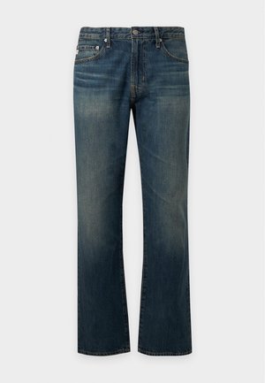 Jonge man die een simpele witte T-shirt, donkerblauwe jeans en witte sneakers draagt, staand tegen een eenvoudige lichte achtergrond.