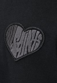 Prenda negra con un logotipo de corazón bordado texturizado en gris oscuro, adornada con rayas verticales y un fondo de tela suave.