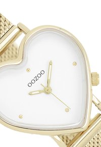 Montre en forme de cœur doré avec un cadran blanc, des accents dorés et un bracelet en maille. Présente un texte de marque noir et trois aiguilles pour l'affichage de l'heure.