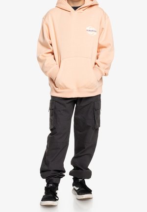 Haut à capuche pêche avec poche avant et logo, associé à un pantalon cargo noir avec poches latérales et chevilles resserrées. Baskets noires et blanches.