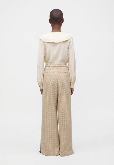 Personne aux cheveux courts portant un chemisier crème à manches longues avec un col à volants et un pantalon large beige à carreaux, debout de dos.