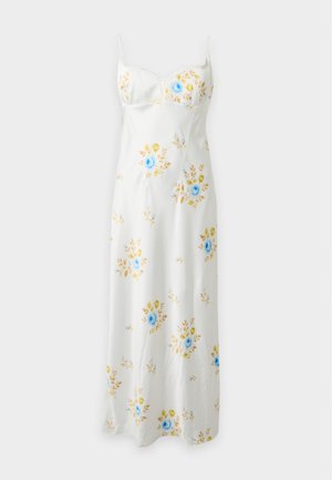 Femme aux longs cheveux foncés portant une robe midi blanche sans manches avec des motifs floraux bleus et jaunes et des chaussures pointues beiges.
