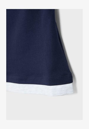 Tessuto blu navy sovrapposto a tessuto grigio chiaro con cuciture visibili ai bordi, che mostra parte dell'orlo di un indumento.