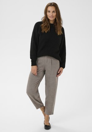 Schwarzer, strukturierter Pullover mit langen Ärmeln, kombiniert mit hellgrauen, lockeren Hosen. Das Model trägt schwarze Ballerinas. Neutrale Hintergrund.