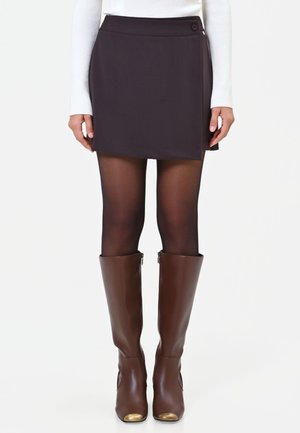 Donna con minigonna marrone scuro, collant neri trasparenti e stivali in pelle marrone al ginocchio con punte dorate, in piedi con le mani rilassate.