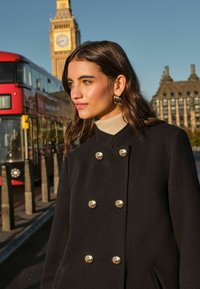 Cappotto nero doppiopetto con bottoni dorati, tessuto testurizzato, indossato sopra un dolcevita beige, ambientato di fronte a uno sfondo cittadino con un autobus rosso.