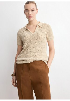 Mujer vistiendo un top de punto beige texturizado de manga corta con cuello y pantalones marrones de pierna ancha y cintura alta, de pie con una mano en el bolsillo.