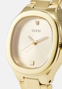 Kuldne metallkell ruudukujulise näo, tekstureeritud heleda kuldse kellapea, kahe peene käe ja väikese ruudukujulise markeriga kell 6. Bränd "GUESS."