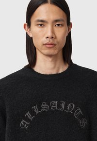 Young man with straight dark hair wears a black fuzzy sweater with dark gray Gothic-style text "TVALSAINES" on a light background.
