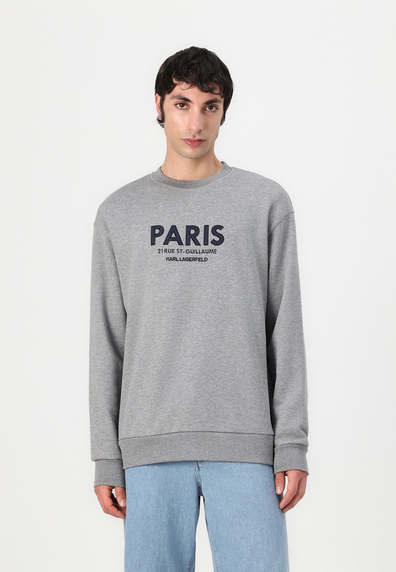 Felpa grigia con maniche lunghe e polsini a coste. Presenta una grande grafica blu "PARIS" e il testo del marchio sottostante. Indossata con jeans azzurri chiari.