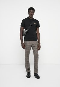 Fekete póló arany logóval, mintás barna nadrág, fekete cipő és egy keresztben viselhető táska. Simára szőtt anyag, testhezálló szabás és kockás dizájn.