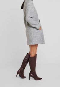 Grauer Wollmischungs-Mantel mit lockerer Passform und Taschen, kombiniert mit glänzenden kastanienbraunen, kniehohen Stiefeln mit Absatz und spitzer Zehenform.