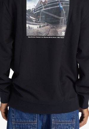 En skateboardåkare utför ett trick på trappor med ledstänger utanför en modern byggnad, tryckt på en svart sweatshirt.
