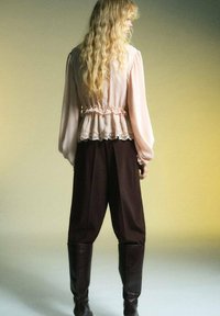 Blouse rose clair avec des manches bouffantes et un bord en dentelle, assortie à un pantalon marron taille haute et des bottes marron jusqu'aux genoux. Textures douces.