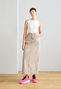 Beige denim maxi skirt with front seam details, paired with a fitted white tank top and bright pink athletic shoes with black laces.