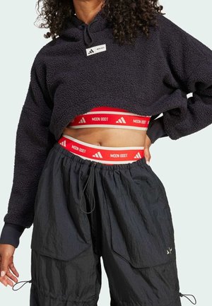 Person iført en kort sort fleece-hoodie, sorte cargobukser og et rødt linning med hvide "Moon Boot"-logoer, der ses på maven.