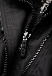 Primer plano de una chaqueta negra con doble cremallera, textura acanalada y logotipo en el tirador de la cremallera. Los dientes de la cremallera son metálicos con acentos negros.