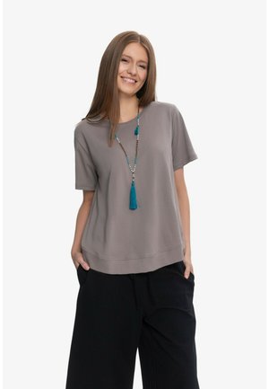 Uśmiechnięta kobieta o długich brązowych włosach nosi luźny szary top z krótkim rękawem, czarne spodnie z szerokimi nogawkami oraz niebieski naszyjnik z koralików z chwostem.