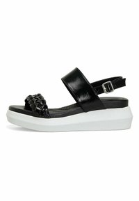 Sandalias planas negras con una correa trenzada, acabado brillante, correa ajustable en el tobillo y suela de goma. Base elevada blanca y plantilla texturizada.