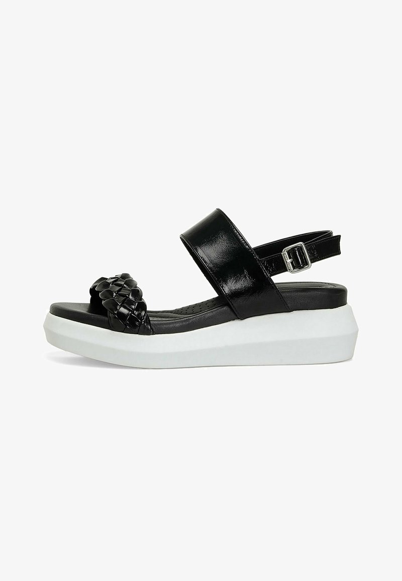 Black platform sandals with a woven strap, glossy finish, adjustable ankle strap, and rubber sole. White elevated base and textured footbed.