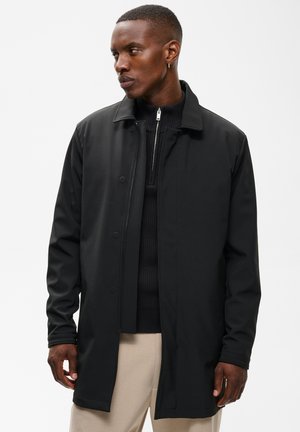 Homme portant un manteau noir par-dessus un pull zippé noir et un pantalon beige, regardant vers sa gauche sur un fond clair uni.