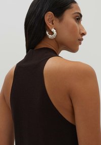 Top in maglia senza maniche di colore nero con un alto colletto, caratterizzato da un design racerback. Orecchini a cerchio in argento con texture visibili all'orecchio sinistro.
