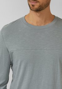 Graues Langarmshirt mit Rundhalsausschnitt, das eine glatte Textur, dezente Nahtdetails über der Brust und einen gerippten Kragen aufweist.