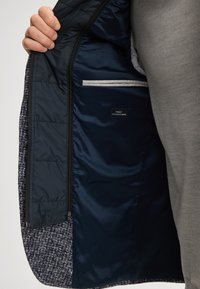 Giacca grigia con tessuto esterno a motivo, fodera in raso blu e tasca laterale con bordo bianco a contrasto ed etichetta.