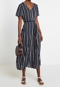 Abito midi a righe blu navy con maniche corte, scollatura a V, bottoni frontali, una cintura in vita e tessuto leggero; abbinato a una borsa rotonda.