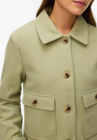 Femme portant un manteau vert olive clair avec de grands boutons et deux poches à rabat boutonnées à l'avant.