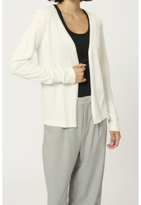 Cardigan bianco a maniche lunghe in morbido tessuto leggero, con scollatura rotonda e apertura davanti. Abbinato a pantaloni grigi chiari, larghi.