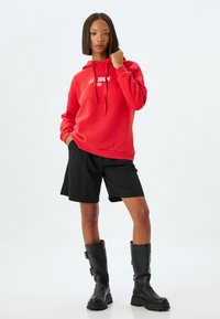 Röd hoodie med vit text, svarta shorts och svarta knähöga stövlar. Hoodien har dragsko och en avslappnad passform.
