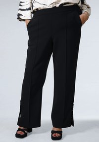 Black wide-leg trousers with a smooth texture, featuring side pockets and decorative buttons at the hem. Worn with platform sandals.