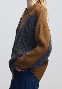 Ylisuuri neulepusero ruskeana ja tummanharmaana, jossa tummalla puolella teksturoitu kaapelikuosi ja resorireunukset.