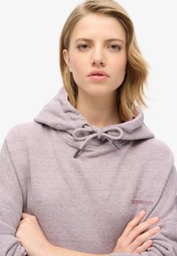 Jasnofioletowa bluza z kapturem z sznurkiem, o miękkiej teksturze i detalu logo na klatce piersiowej. Dopasowany kaptur i luźny krój.