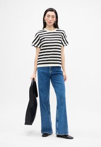 Tricou cu mânecă scurtă în dungi alb-negru, combinat cu blugi evazați din denim albastru. Modelul ține o geantă de mână neagră. Design simplu, croială casual.
