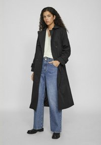 Palton negru tip trench cu nasturi dublu-efecta, blugi albaștri cu picioare largi și cizme negre până la glezne. Modelul stă în fața unui fundal simplu gri.