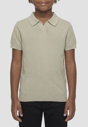 Bambino che indossa una polo beige a maniche corte con texture e pantaloni neri, in piedi su uno sfondo chiaro e uniforme.