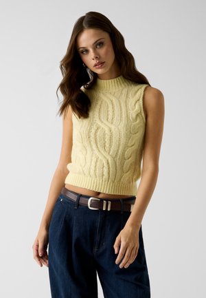 Giovane donna con lunghi capelli castani che indossa un gilet lavorato a maglia giallo senza maniche e pantaloni blu scuro a vita alta con una cintura marrone.