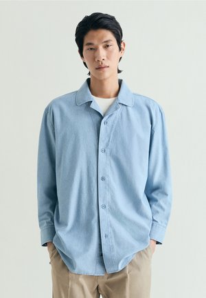 Joven con cabello negro vistiendo una camisa azul claro abotonada sobre una camiseta blanca y pantalones beige, de pie con las manos en los bolsillos.