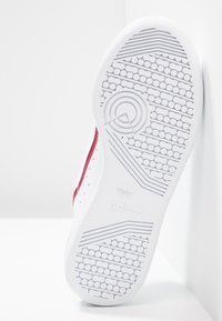 Sneaker bianco con suola in gomma, accenti rossi e blu navy, dettagli perforati e modelli texturizzati sulla suola. Logo Adidas visibile.