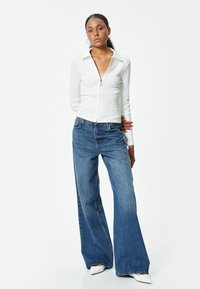 Maglia bianca a coste con zip e colletto, abbinata a jeans a gamba larga blu. La modella indossa scarpe bianche con punta a punta. Sfondo semplice.