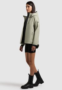 Femme debout portant une veste à capuche gris clair, un short noir et des bottes noires mi-mollet sur un fond clair uni.