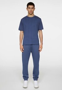 Sininen urheilupaita ja pehmeästä kankaasta valmistetut verkkarit. Asussa on pyöreä pääntie, rento istuvuus ja kiristysnyörit vyötäröllä. Valkoiset lenkkarit.