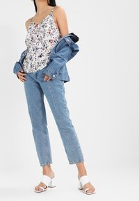 Donna che indossa un top floreale, giacca di jeans leggera blu chiaro drappeggiata sulle spalle, jeans blue straight-leg e sandali con tacco bianchi.