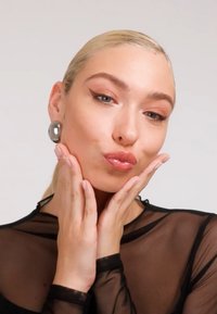 Svart gjennomsiktig topp med høy hals, store metalliske øredobber og naturlig sminke; hender som holder kinnene, fremhever ansiktstrekk.