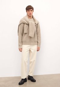 Suéter beige acanalado llevado sobre un suéter liviano, combinado con pantalones crema y zapatos negros de cordones. Tejidos suaves y texturizados, con un ajuste casual.