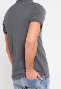 Šedé polo tričko s límečkem, krátkými rukávy a mírným zakřivením na spodním okraji. Tkanina se zdá být měkká s hladkým povrchem.